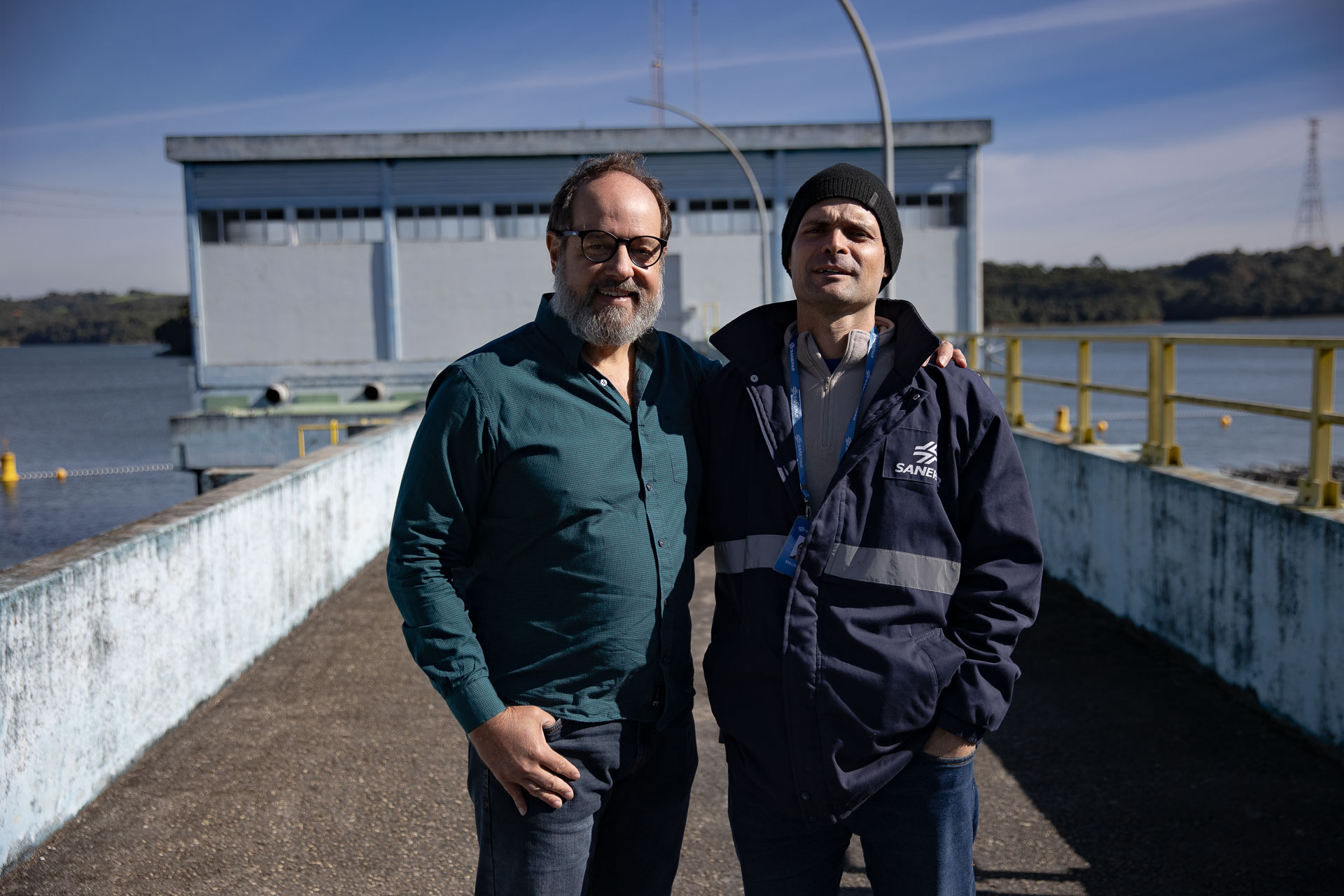 Dois homens posam abraçados de lado na passarela externa da estação de captação do Reservatório do Passaúna. O homem à esquerda usa óculos, barba e camisa verde, e o homem à direita veste uma jaqueta escura com a logomarca da Sanepar e um gorro preto, sob a luz do sol.