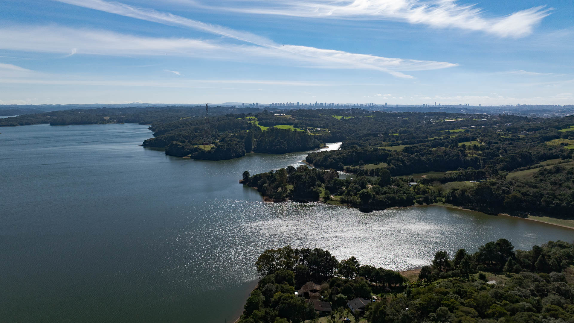 Vista aérea da Represa do Passaúna cercada por áreas verdes, com a silhueta de prédios no horizonte sob céu claro.