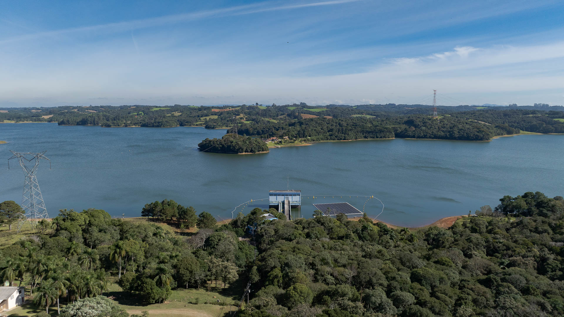 Vista aérea da estação de captação de água da Sanepar no Reservatório do Passaúna, cercada por densa vegetação nativa, com placas solares flutuantes na água e torres de transmissão de energia ao fundo sob céu azul.