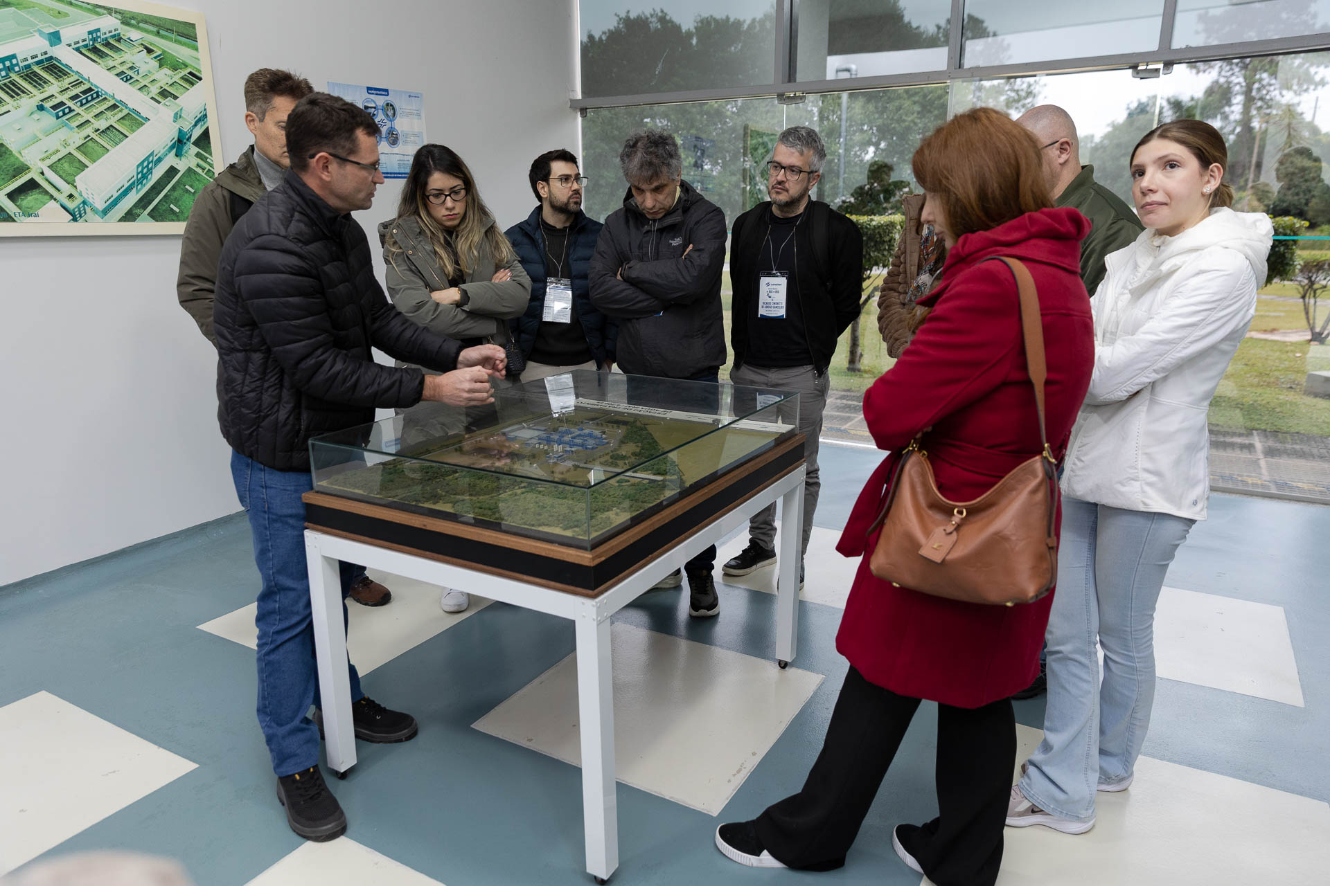 Grupo de visitantes observa uma maquete física detalhada de uma estação de tratamento de água sob proteção de vidro, enquanto um técnico da Sanepar faz explicações apontando para a estrutura