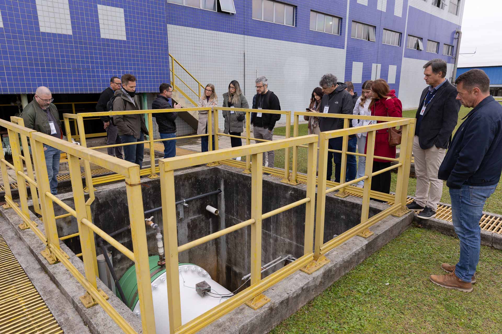 Visitantes apoiados em um guarda-corpo amarelo observam o interior de um tanque profundo de concreto com tubulações nas instalações da Sanepar