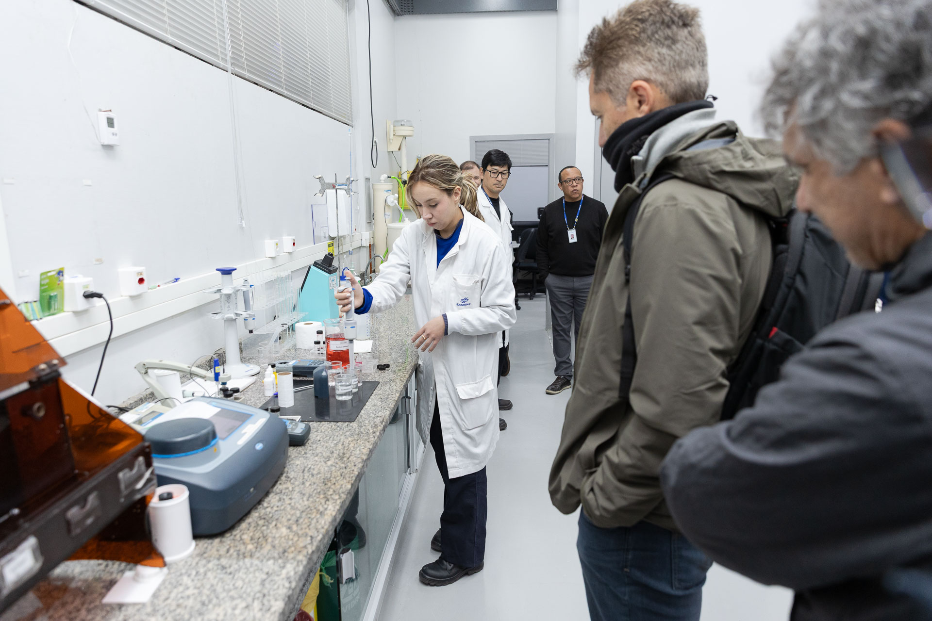 Profissional de jaleco branco utiliza uma pipeta em bancada de laboratório de análises de água, sob o olhar atento de integrantes da comitiva