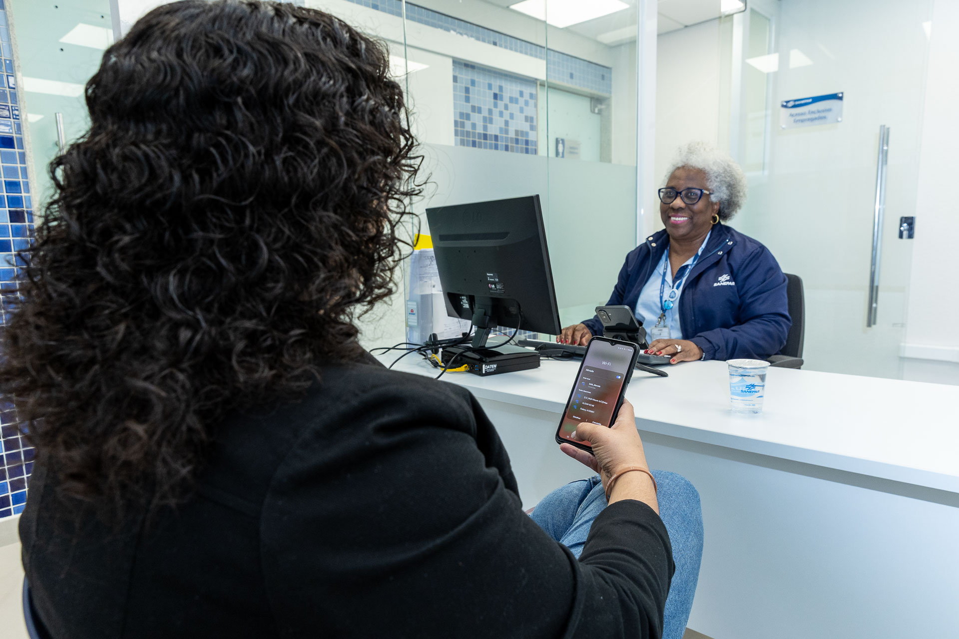 clientes está no atendimento da sanepar e está usando o celular. cliente tem cabelos cacheados e aparece de costas, enquanto uma atendente sorri na sua frente