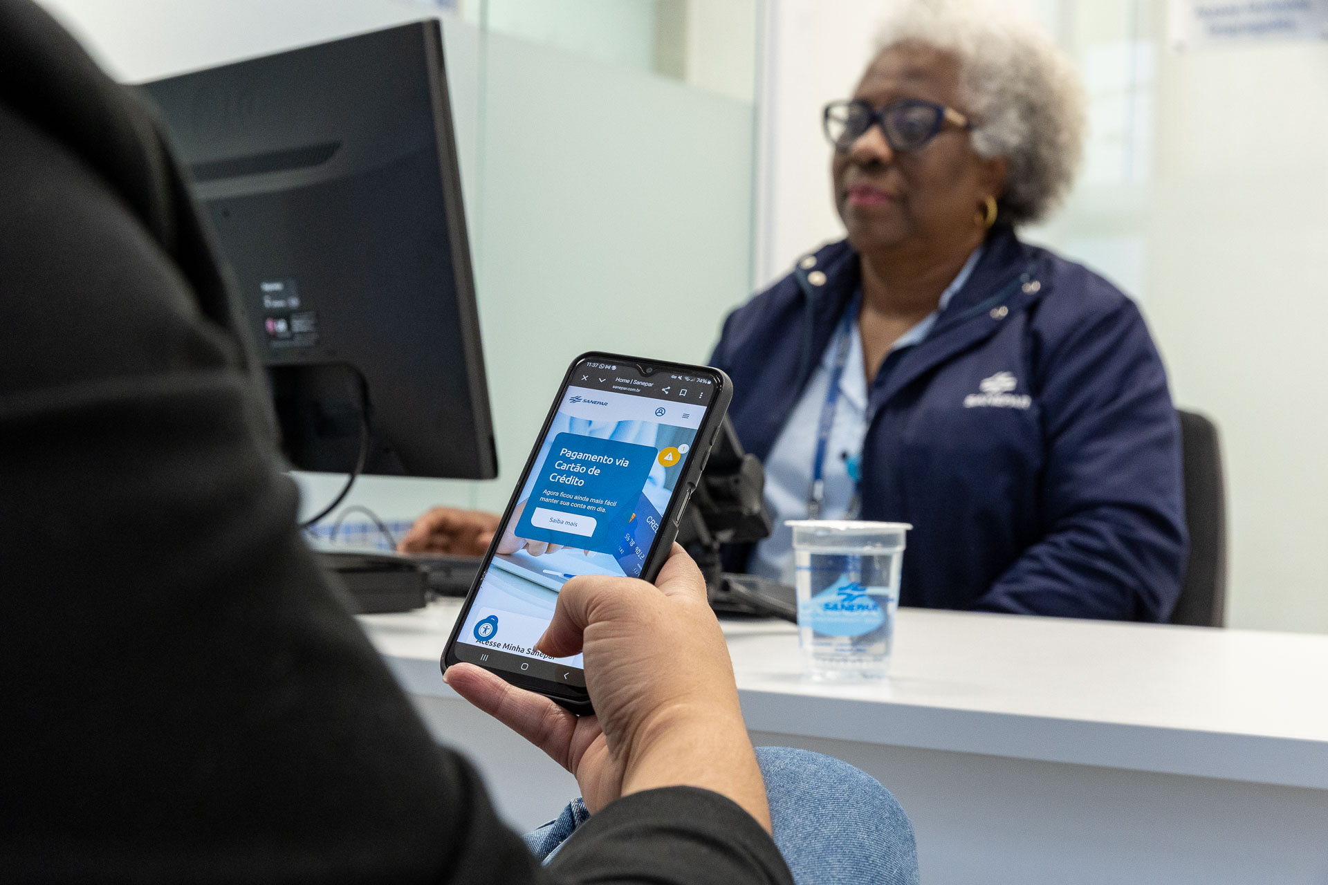 clientes está no atendimento da sanepar e está usando o celular. uma mulher negra e de cabelos brancos é a atendente