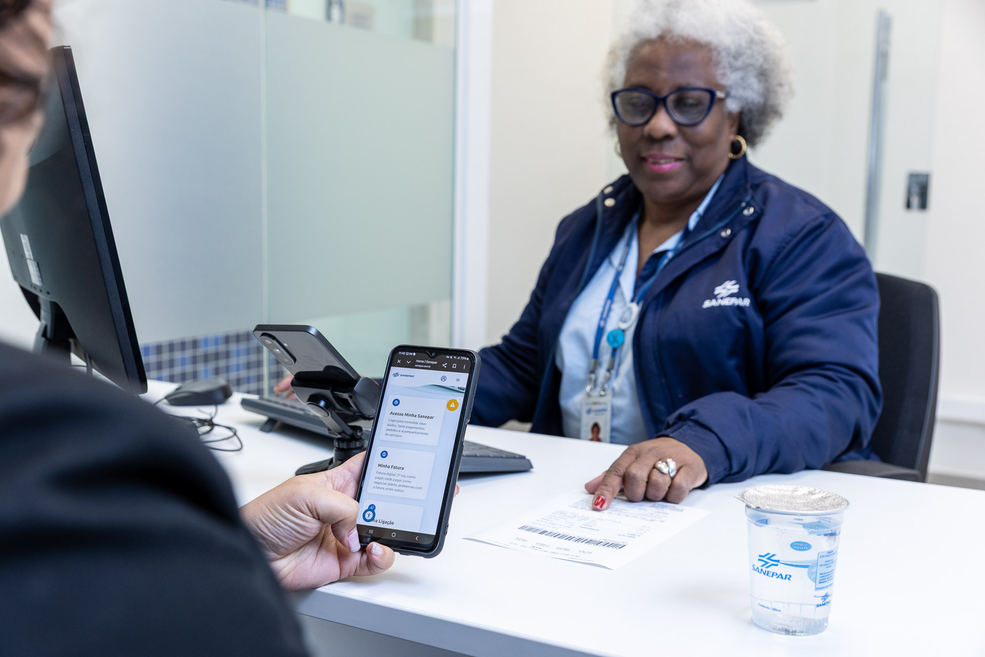 clientes está no atendimento da sanepar e está usando o celular. uma mulher negra e de cabelos brancos é a atendente