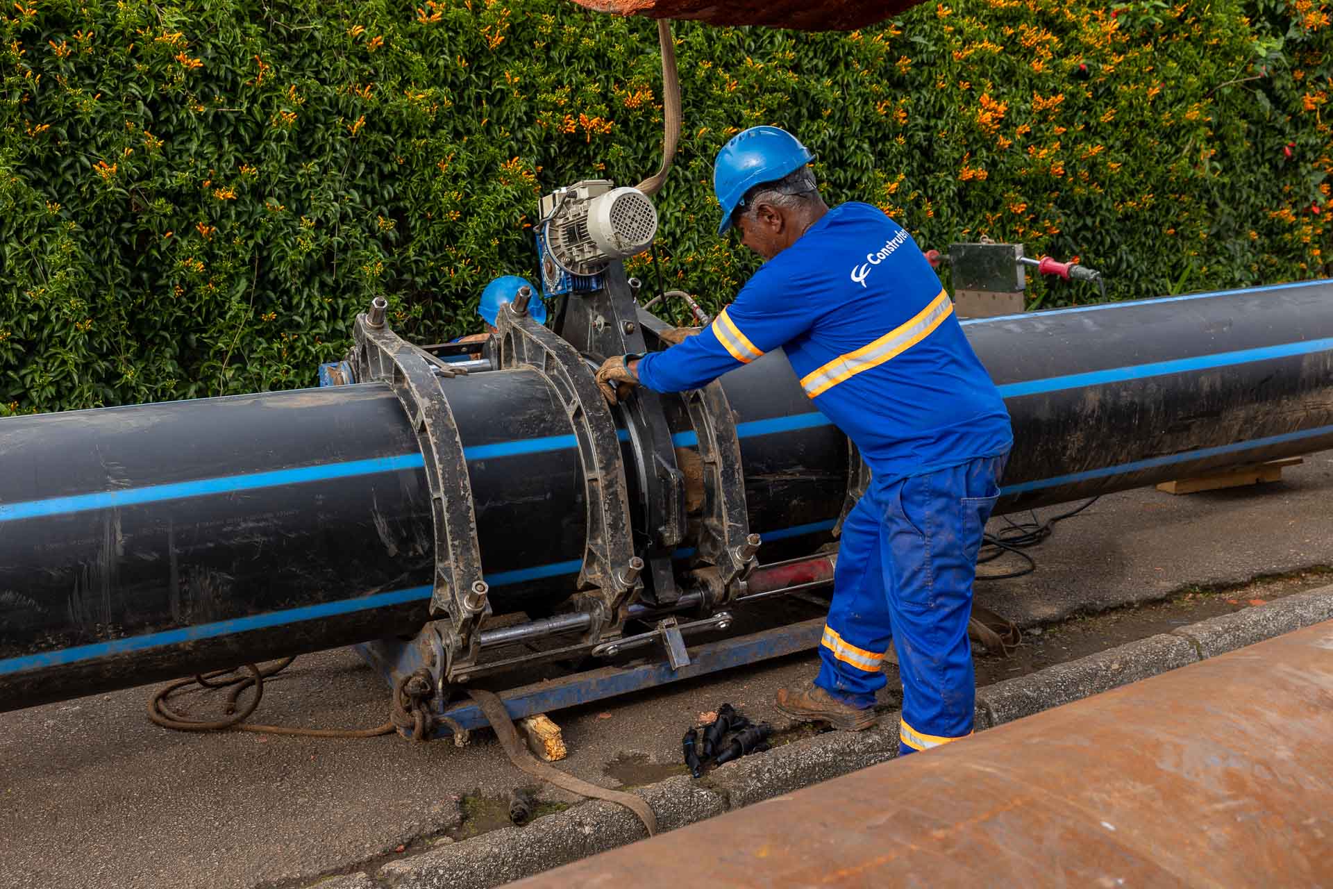 Operário da Sanepar ajustando uma máquina de fixação e solda térmica instalada ao redor de uma grande tubulação de água preta com listra azul.