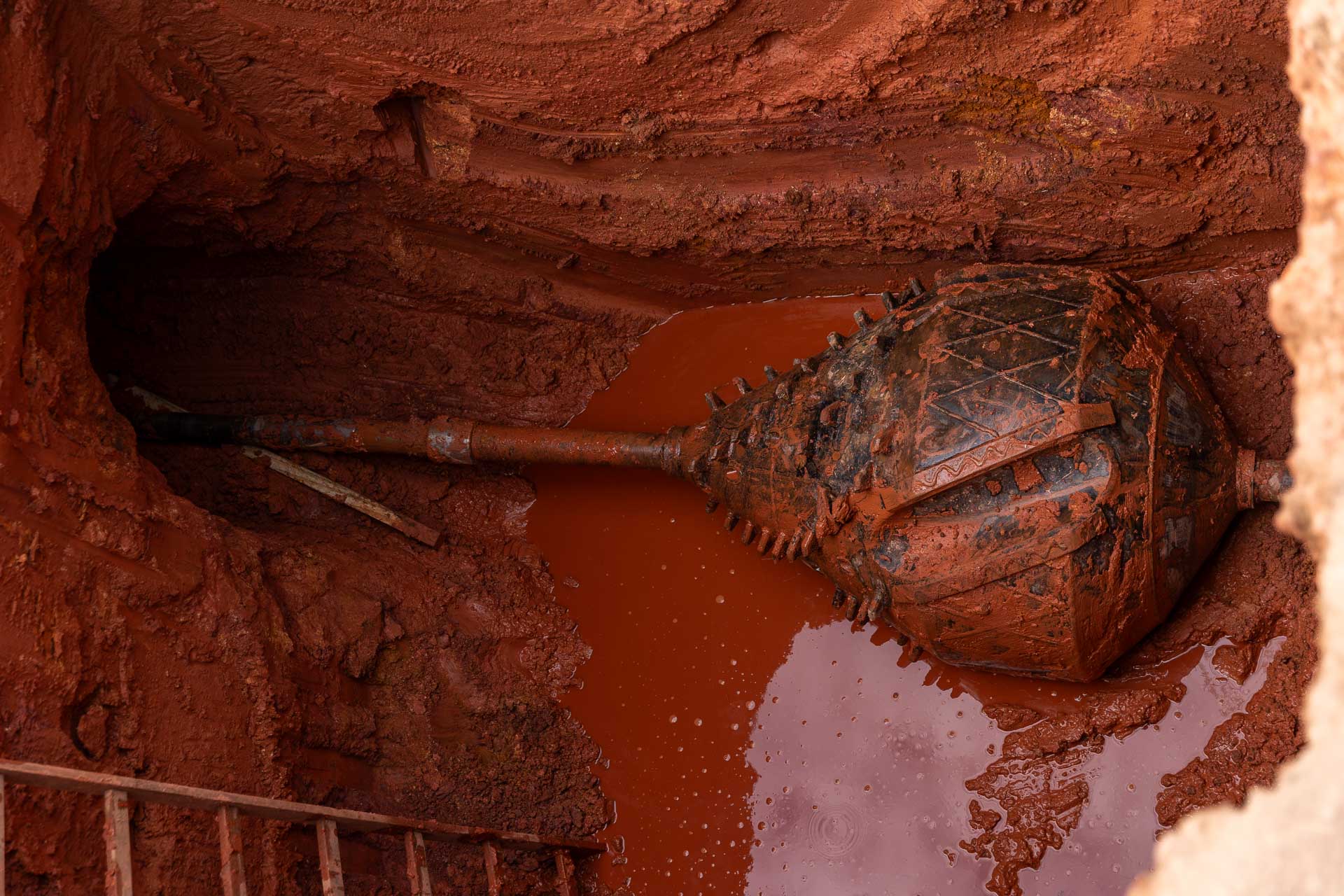 Grande broca de perfuração de formato oval suja de lama acoplada a um cano, posicionada dentro de uma escavação profunda de terra vermelha.