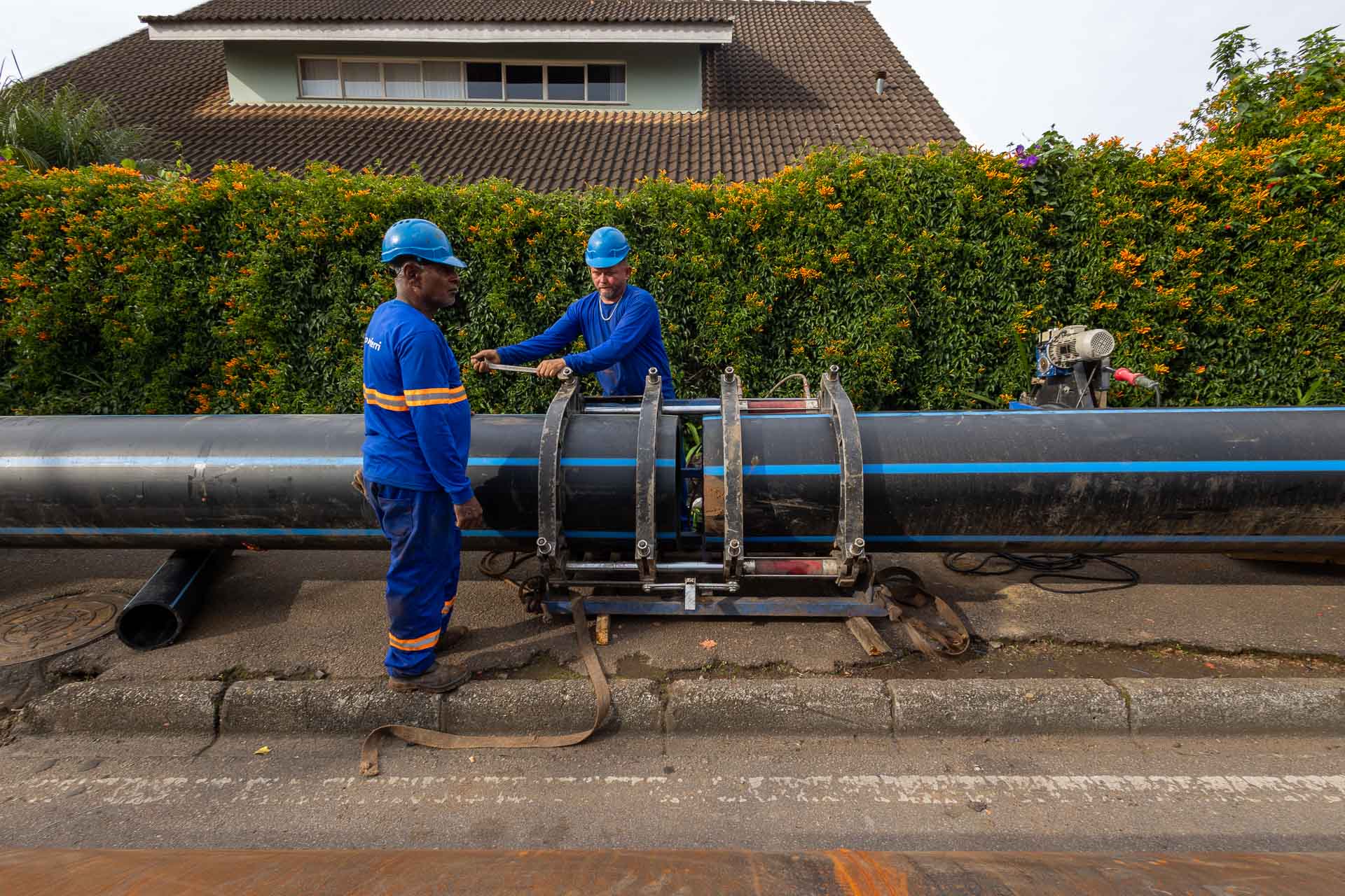 Dois operários de uniforme azul e capacete operando um equipamento alinhador para conectar duas grandes tubulações pretas com listras azuis na rua.