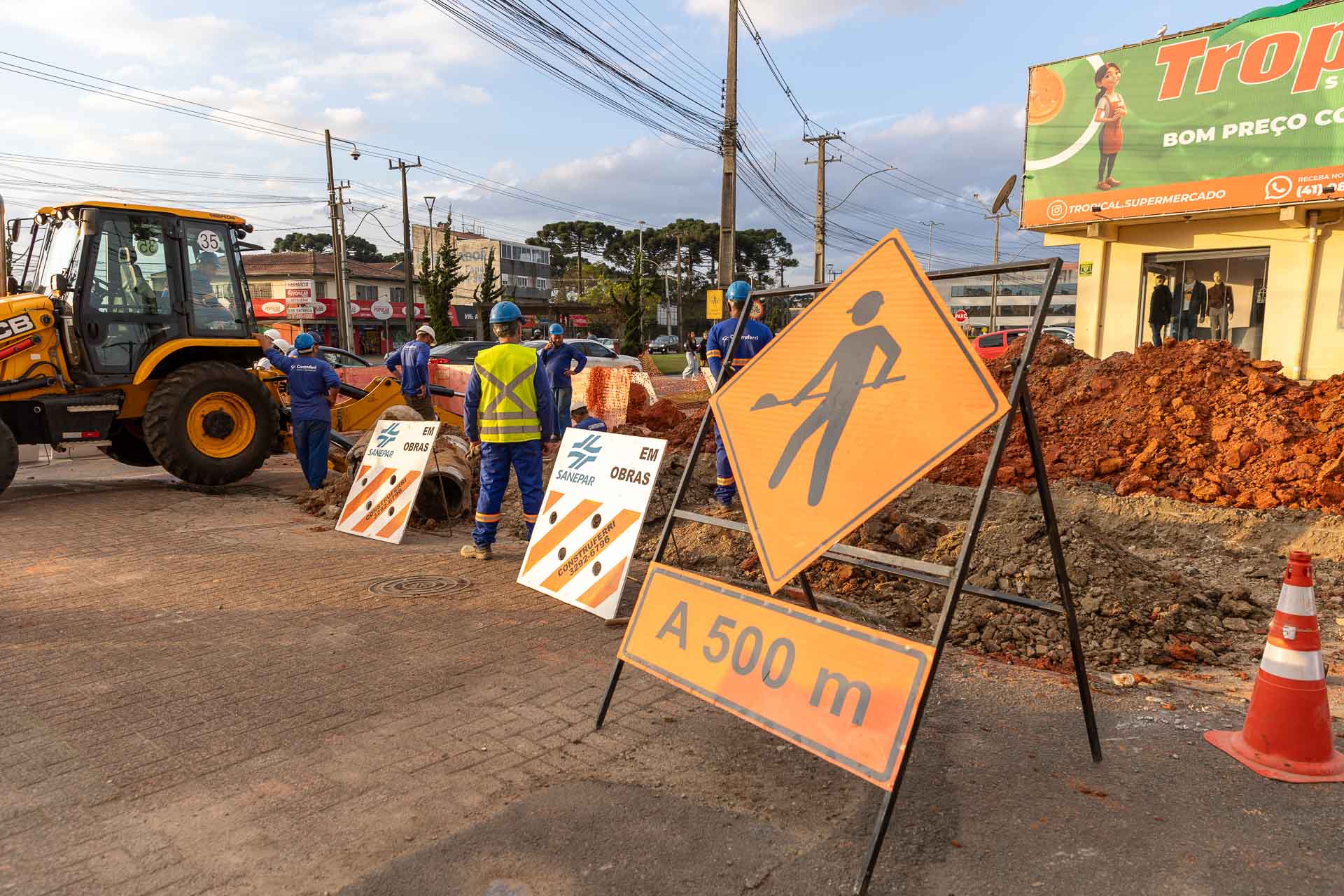 Canteiro de obras com placa laranja indicando homens trabalhando e cavaletes da Sanepar em primeiro plano, com operários e uma retroescavadeira ao fundo.