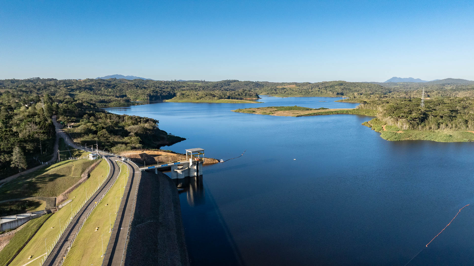 Vista panorâmica da Barragem do Miringuava com amplo reservatório de água cercado por vegetação e morros ao fundo sob céu limpo