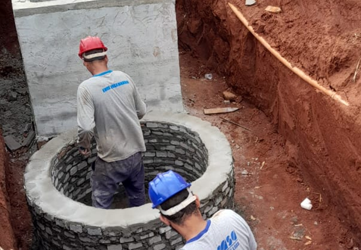 dois homens vestindo roupa cinza e capacete fazem obra civil com tijolos e cimento