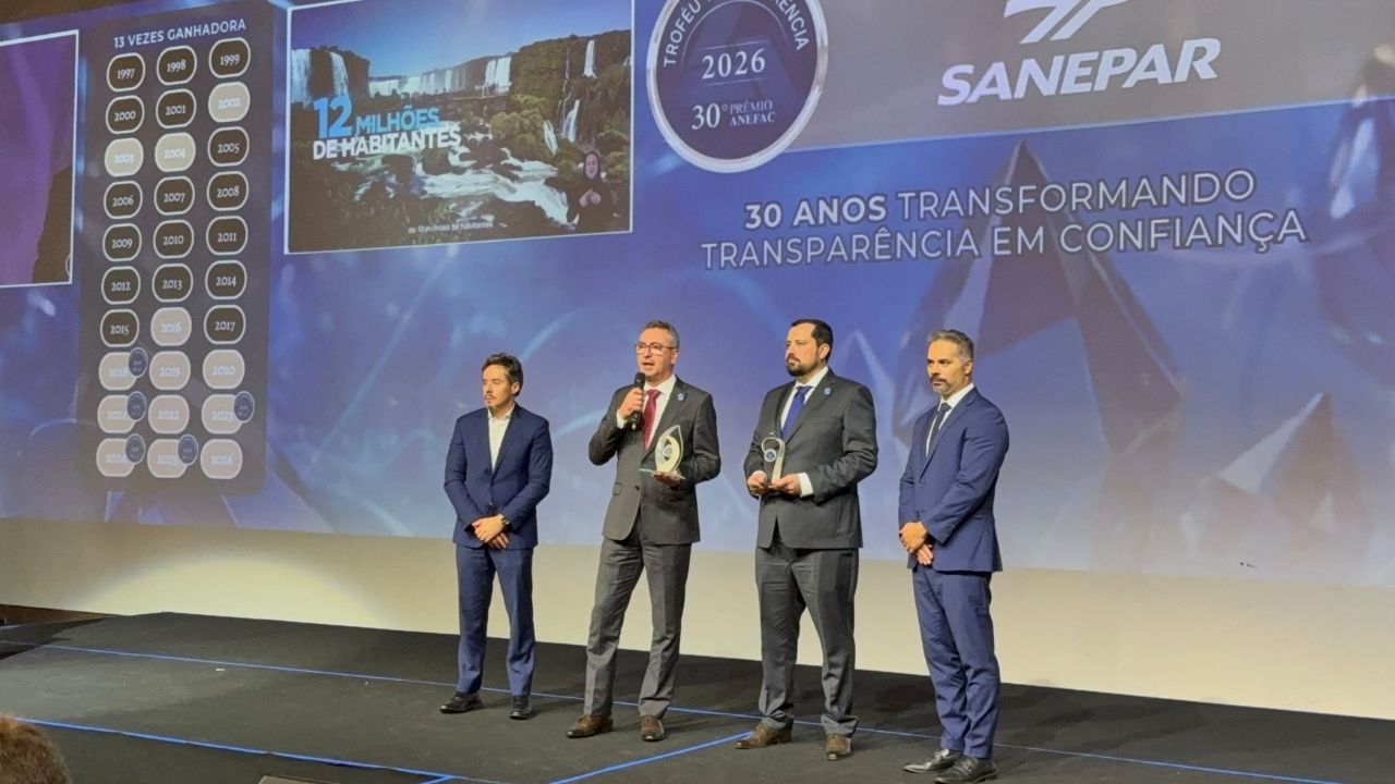 Quatro homens em um palco, sendo que um deles fala ao microfone segurando um troféu, com um telão ao fundo exibindo a marca Sanepar, o selo do Troféu Transparência 2026 e a frase "30 anos transformando transparência em confiança".