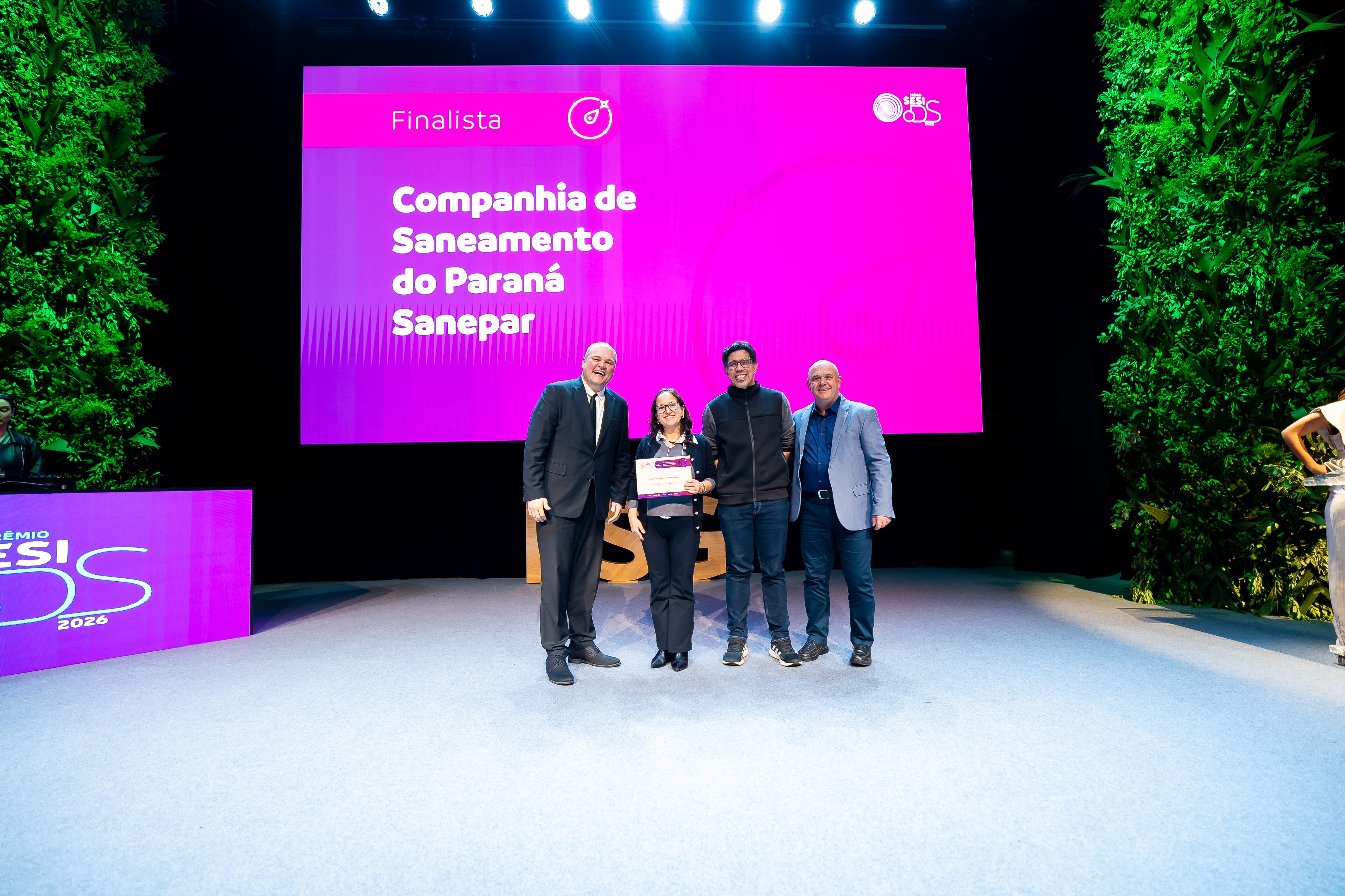 Três pessoas posam sorridentes no palco segurando um certificado. Ao fundo, o telão rosa do evento apresenta o texto "Finalista Companhia de Saneamento do Paraná Sanepar".