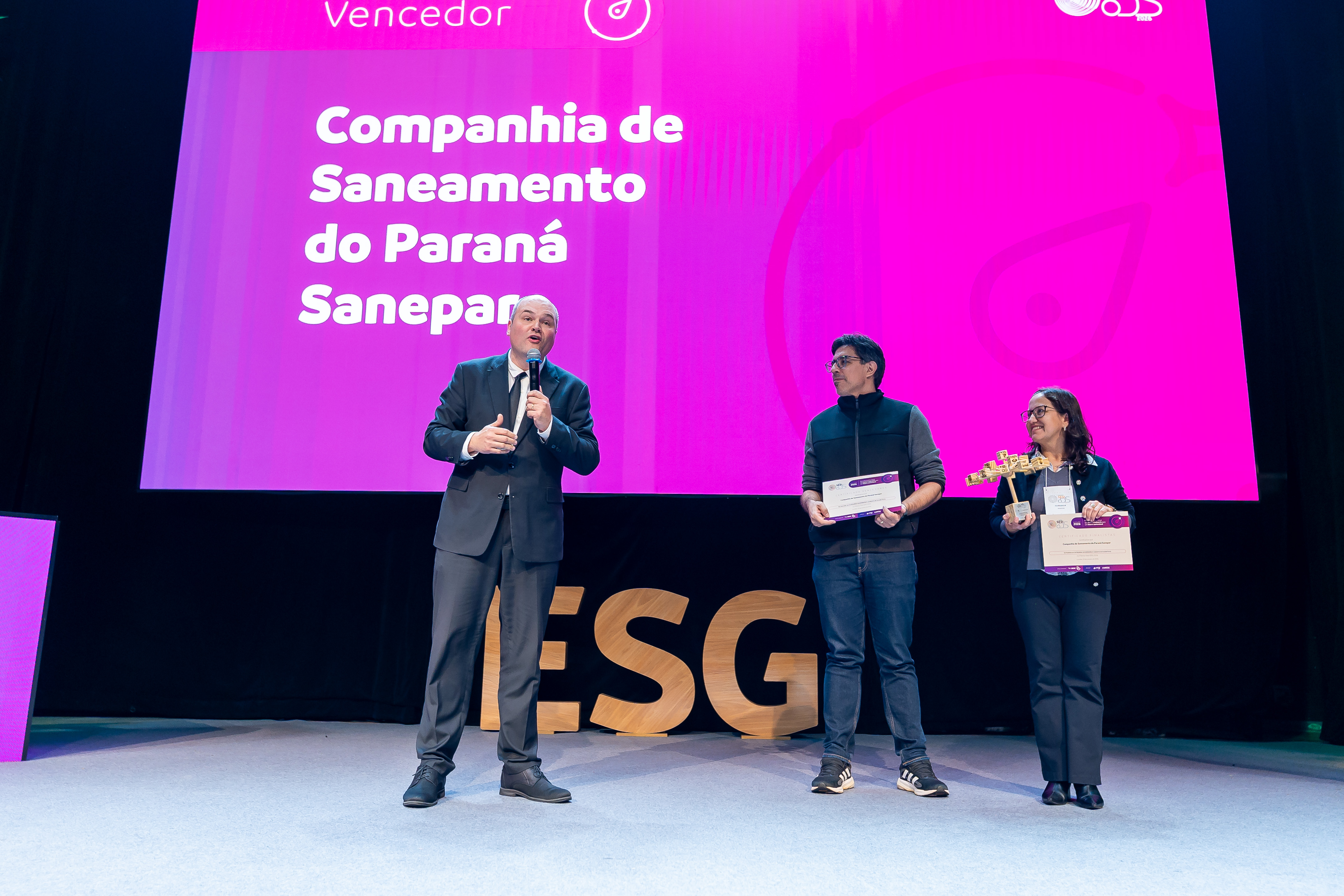 Três representantes no palco do Prêmio Sesi ODS 2026. Um homem à esquerda fala ao microfone, enquanto um homem e uma mulher à direita seguram um certificado e um troféu. O telão rosa ao fundo destaca o texto "Vencedor Companhia de Saneamento do Paraná Sanepar".