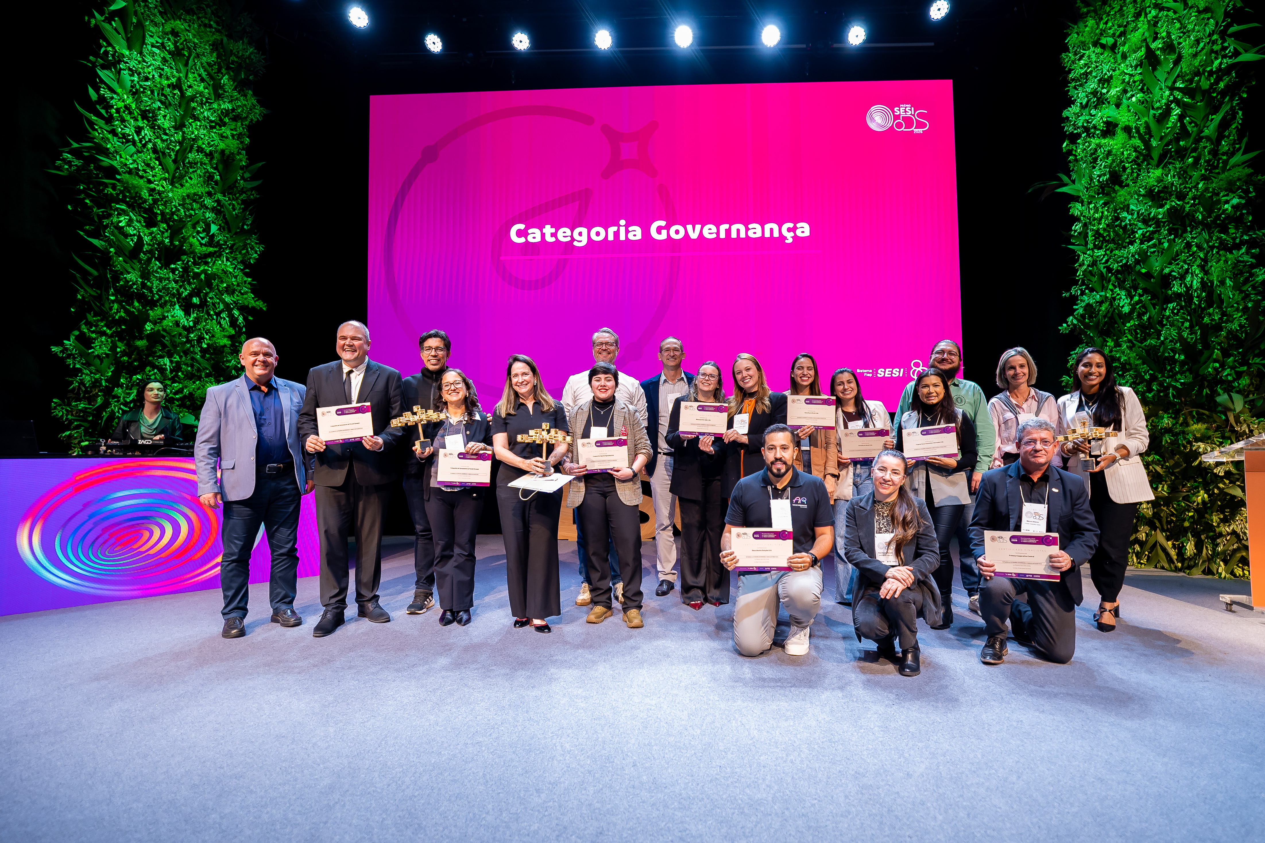 Grupo de pessoas sorridentes posa no palco segurando troféus e certificados. Ao fundo, um grande telão rosa exibe a logomarca do Prêmio Sesi ODS 2026 e o texto "Categoria Governança".