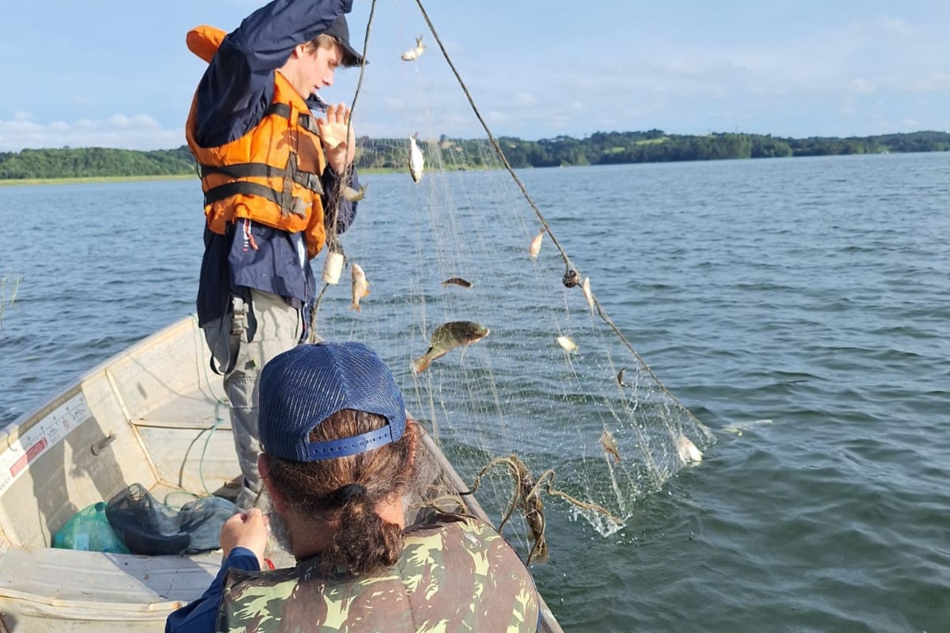 Dois pesquisadores em um barco de alumínio recolhem uma rede de pesca com pequenos lambaris-miúdos e outros peixes, durante um estudo ambiental em um reservatório de água sob céu claro.