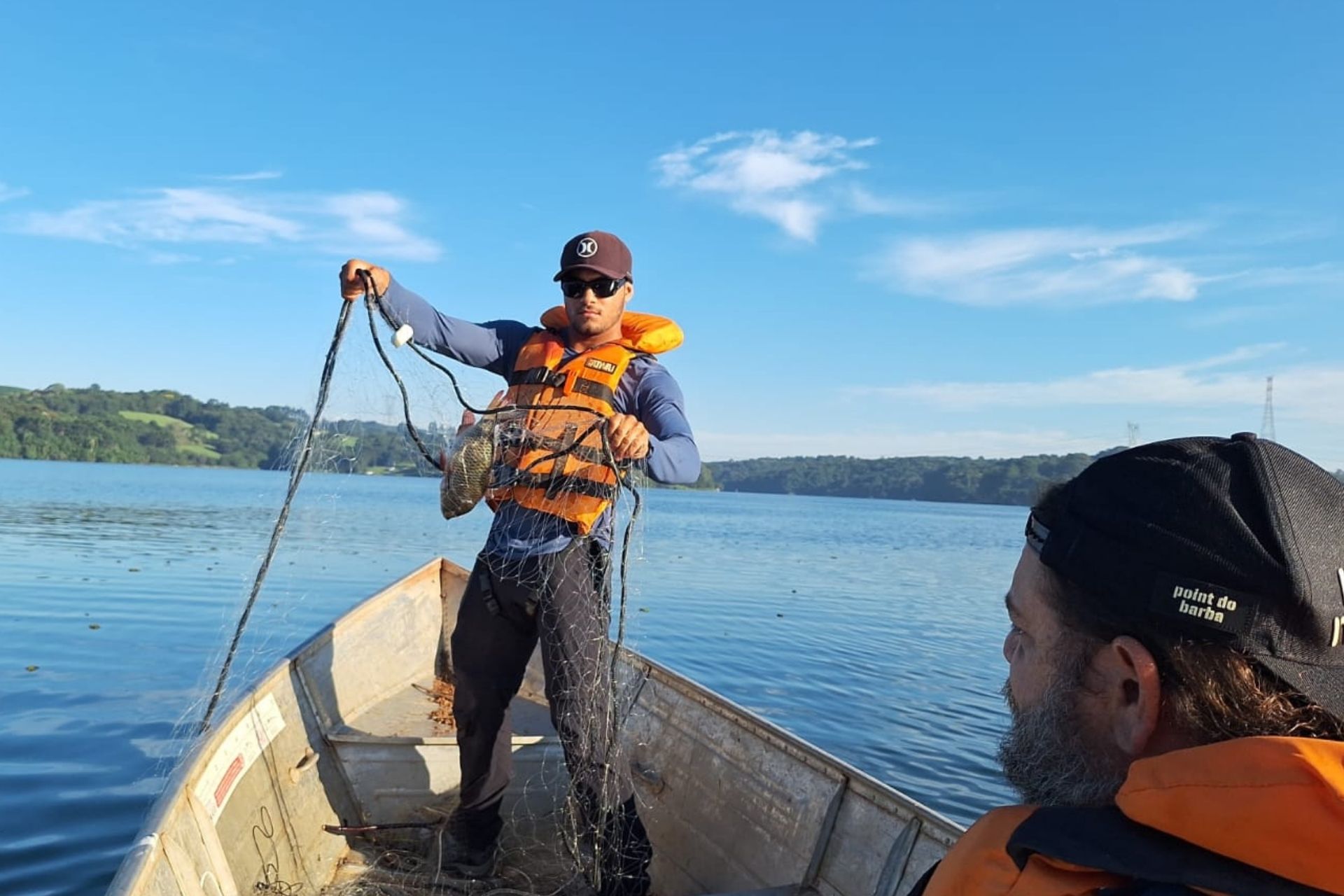 Pesquisador em pé na proa de um barco de alumínio segura uma rede de pesca com um peixe preso, enquanto outro homem em primeiro plano observa a atividade em um reservatório de água sob céu azul. Crédito: Matheus Oliveira Freitas