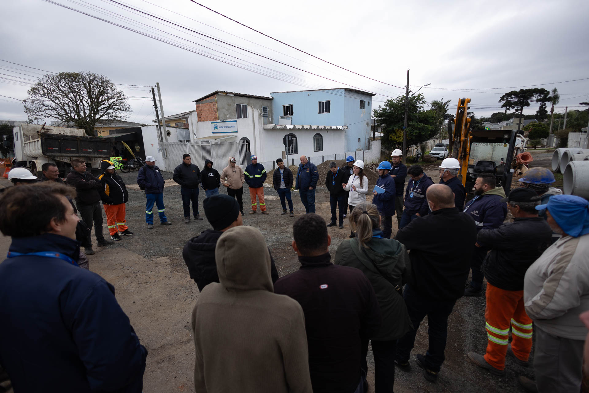 Vista ampliada de uma reunião técnica ao ar livre em uma rua residencial, onde trabalhadores e representantes da Sanepar e da prefeitura formam uma roda para acompanhar as orientações sobre a obra