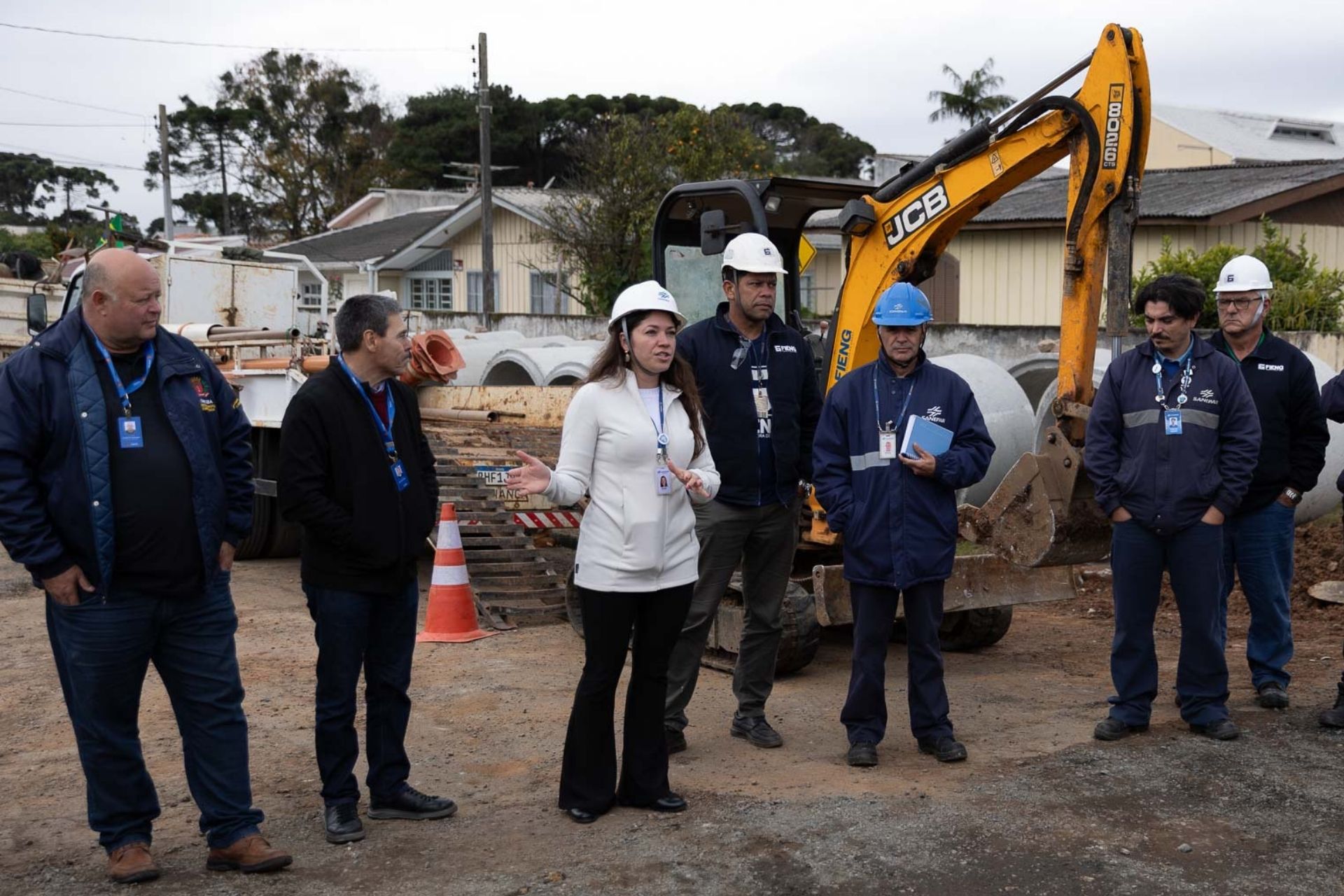 Representante da Sanepar com capacete e casaco brancos gesticula enquanto fala com equipe técnica e trabalhadores em um canteiro de obras, tendo ao fundo tubulações de concreto e uma escavadeira amarela.