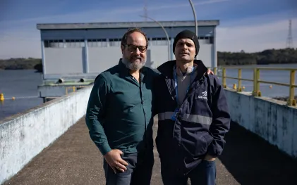 Dois homens posam abraçados de lado na passarela externa da estação de captação do Reservatório do Passaúna. O homem à esquerda usa óculos, barba e camisa verde, e o homem à direita veste uma jaqueta escura com a logomarca da Sanepar e um gorro preto, sob a luz do sol.