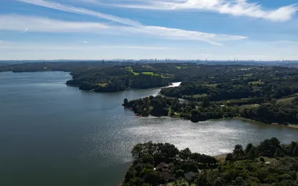 Vista aérea da Represa do Passaúna cercada por áreas verdes, com a silhueta de prédios no horizonte sob céu claro.