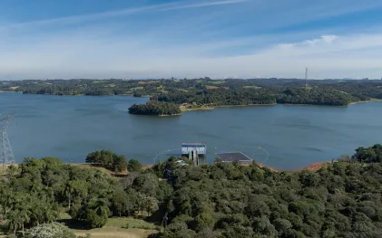 Vista aérea da estação de captação de água da Sanepar no Reservatório do Passaúna, cercada por densa vegetação nativa, com placas solares flutuantes na água e torres de transmissão de energia ao fundo sob céu azul.