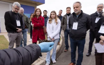 Primeiro plano focado em uma mão com luva azul segurando um pedaço de lodo desidratado, com o grupo de visitantes ao fundo prestando atenção