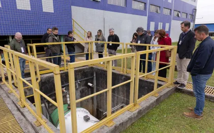 Visitantes apoiados em um guarda-corpo amarelo observam o interior de um tanque profundo de concreto com tubulações nas instalações da Sanepar