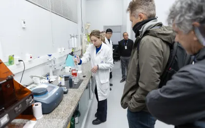 Profissional de jaleco branco utiliza uma pipeta em bancada de laboratório de análises de água, sob o olhar atento de integrantes da comitiva