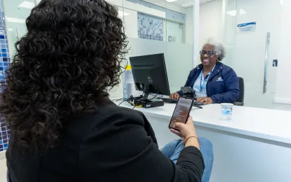 clientes está no atendimento da sanepar e está usando o celular. cliente tem cabelos cacheados e aparece de costas, enquanto uma atendente sorri na sua frente