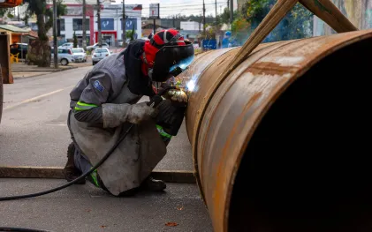 Soldador utilizando capa de proteção e máscara enquanto faz a solda de uma grande tubulação metálica no meio-fio da rua, gerando faíscas brilhantes.