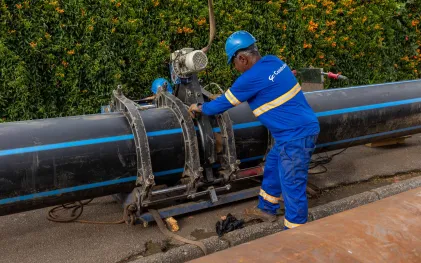 Operário da Sanepar ajustando uma máquina de fixação e solda térmica instalada ao redor de uma grande tubulação de água preta com listra azul.