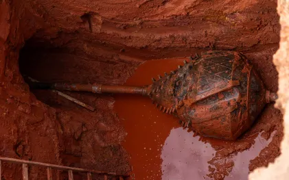 Grande broca de perfuração de formato oval suja de lama acoplada a um cano, posicionada dentro de uma escavação profunda de terra vermelha.