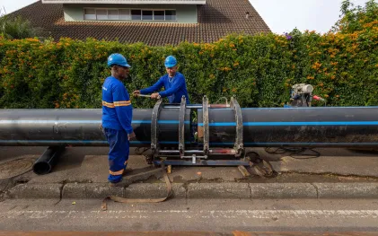 Dois operários de uniforme azul e capacete operando um equipamento alinhador para conectar duas grandes tubulações pretas com listras azuis na rua.