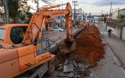 Escavadeira laranja posicionada ao lado de uma extensa vala de terra vermelha em uma rua, com operários da Sanepar e outra máquina trabalhando ao fundo.
