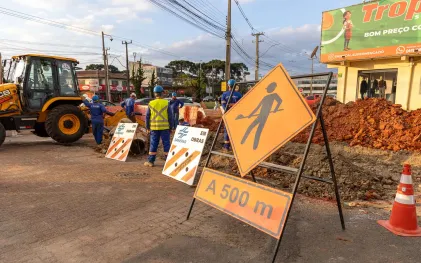 Canteiro de obras com placa laranja indicando homens trabalhando e cavaletes da Sanepar em primeiro plano, com operários e uma retroescavadeira ao fundo.