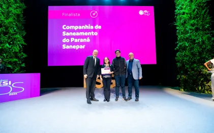 Três pessoas posam sorridentes no palco segurando um certificado. Ao fundo, o telão rosa do evento apresenta o texto "Finalista Companhia de Saneamento do Paraná Sanepar".
