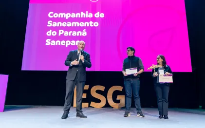 Três representantes no palco do Prêmio Sesi ODS 2026. Um homem à esquerda fala ao microfone, enquanto um homem e uma mulher à direita seguram um certificado e um troféu. O telão rosa ao fundo destaca o texto "Vencedor Companhia de Saneamento do Paraná Sanepar".