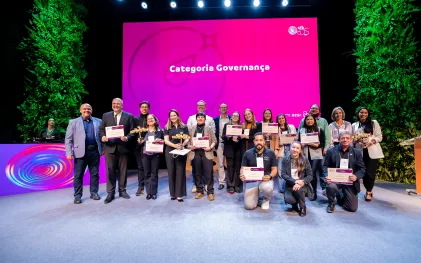 Grupo de pessoas sorridentes posa no palco segurando troféus e certificados. Ao fundo, um grande telão rosa exibe a logomarca do Prêmio Sesi ODS 2026 e o texto "Categoria Governança".