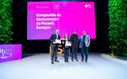 Quatro pessoas posam juntas no palco. Uma mulher ao centro segura um troféu e um certificado, ladeada por três homens. Ao fundo, o telão rosa do evento exibe o texto "Vencedor Companhia de Saneamento do Paraná Sanepar".