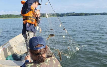Dois pesquisadores em um barco de alumínio recolhem uma rede de pesca com pequenos lambaris-miúdos e outros peixes, durante um estudo ambiental em um reservatório de água sob céu claro.