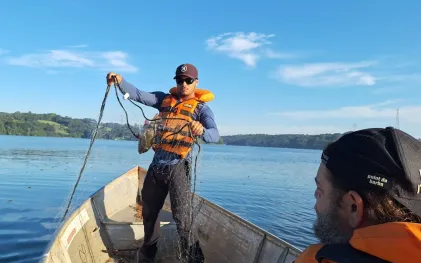 Pesquisador em pé na proa de um barco de alumínio segura uma rede de pesca com um peixe preso, enquanto outro homem em primeiro plano observa a atividade em um reservatório de água sob céu azul. Crédito: Matheus Oliveira Freitas