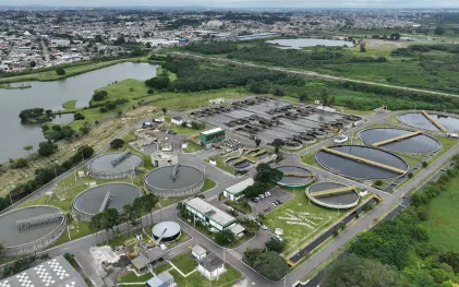Vista aérea da estação de tratamento de esgoto Belém com tanques decantadores circulares e lagoa adjacente, com área urbana ao fundo.