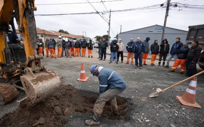 Operário com uniforme sujo de terra trabalha com uma pá em uma escavação no asfalto, ao lado de uma escavadeira, enquanto um grupo de trabalhadores com uniformes coloridos e técnicos da Sanepar observam a atividade