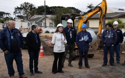 Representante da Sanepar com capacete e casaco brancos gesticula enquanto fala com equipe técnica e trabalhadores em um canteiro de obras, tendo ao fundo tubulações de concreto e uma escavadeira amarela.
