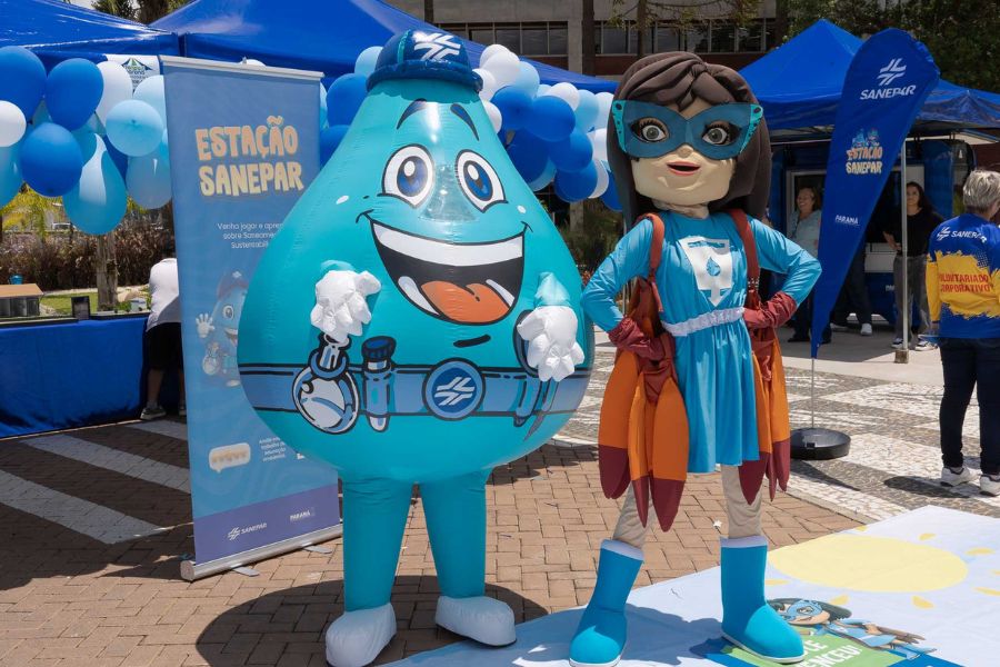 Mascote da Sanepar em formato de gota d'água sorridente e super-heroína com capa e máscara azuis posam lado a lado em evento ao ar livre