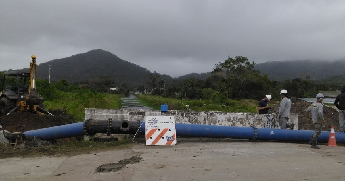 Tubulação azul de grande porte cruza um canal apoiada sobre estrutura de concreto. Operários com capacetes de segurança trabalham na estrutura ao lado de uma retroescavadeira e de uma placa que indica "Em obras Sanepar". Ao fundo, montanhas sob céu nublado.