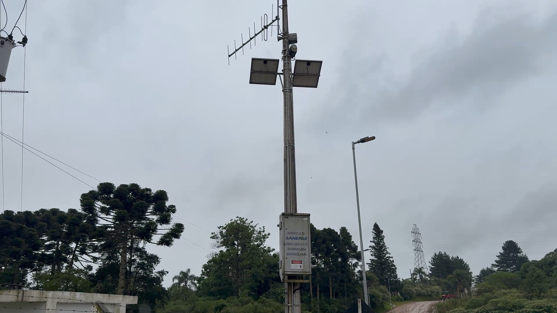 Fotografia em ângulo contrapicado de um poste de concreto que sustenta equipamentos do sistema de alerta da Sanepar, sob um céu nublado e cinzento. Próximo ao topo do poste, há duas placas de energia solar e antenas de transmissão direcionais. Na parte inferior do poste, uma caixa metálica cinza traz inscrições técnicas identificando o equipamento como "REMOTA 01 SANEPAR", acompanhada do telefone de contato 0800 e da indicação "BARRAGEM MIRINGUAVA".