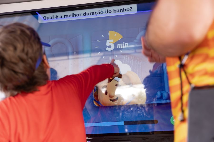 Criança toca em tela interativa que exibe a pergunta "Qual é a melhor duração do banho?" com resposta destacada de "5 min"