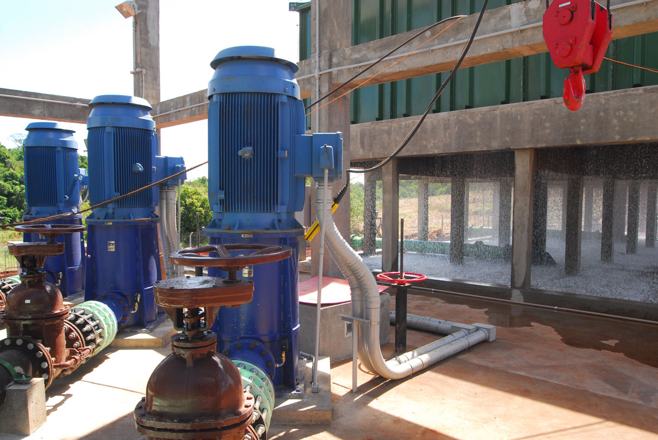 Conjunto de grandes motores elétricos azuis acoplados a tubulações e registros em área operacional. Ao fundo, estrutura de concreto com cortina de água em cascata.